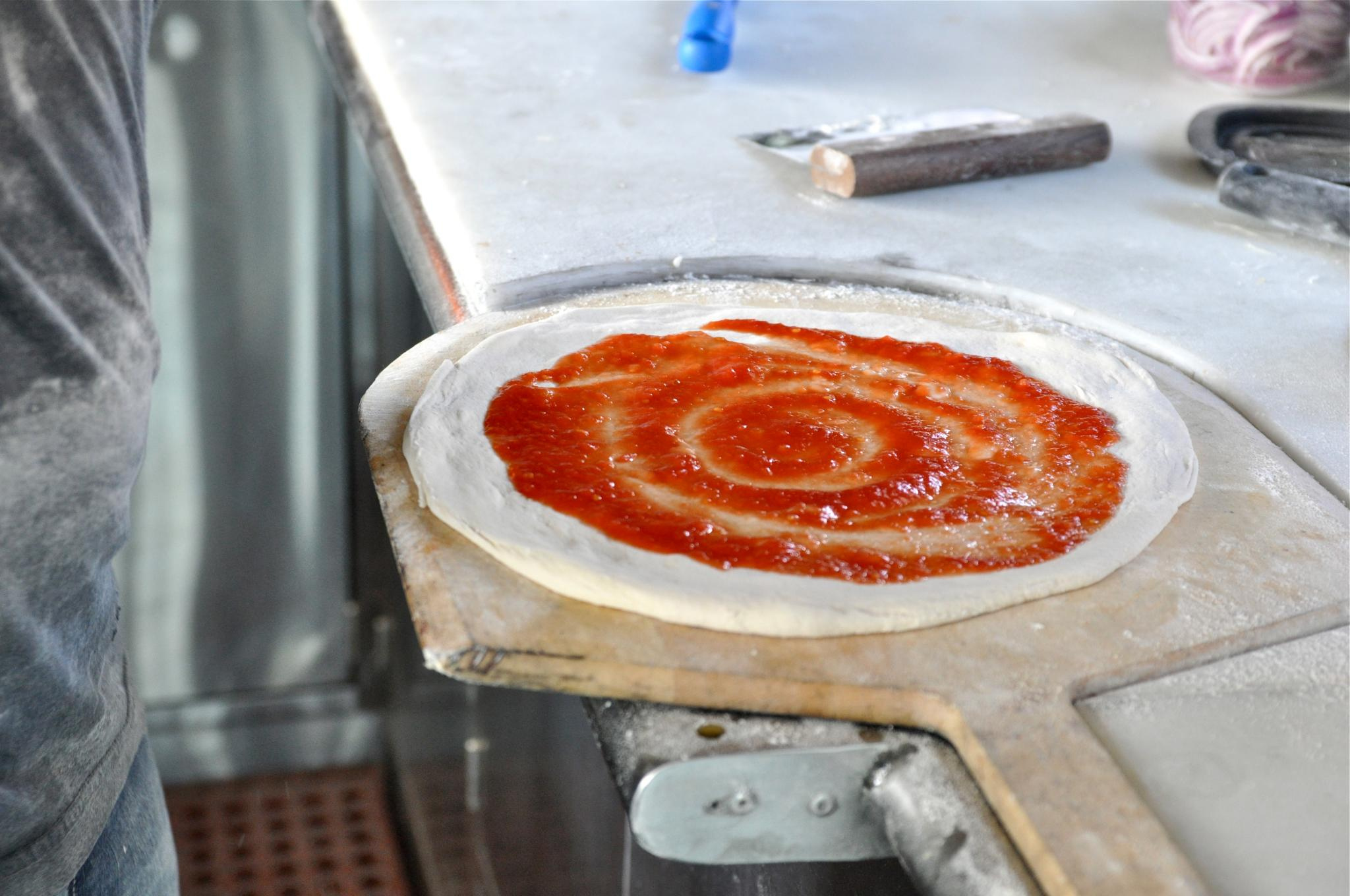 A round-shaped uncooked pizza dough with a spiral of sauce on top. It's lying on a wood pizza peel.